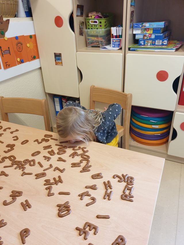 Ein Mädchen versucht Buchstaben aus Russischem Brot vom Tisch zu essen